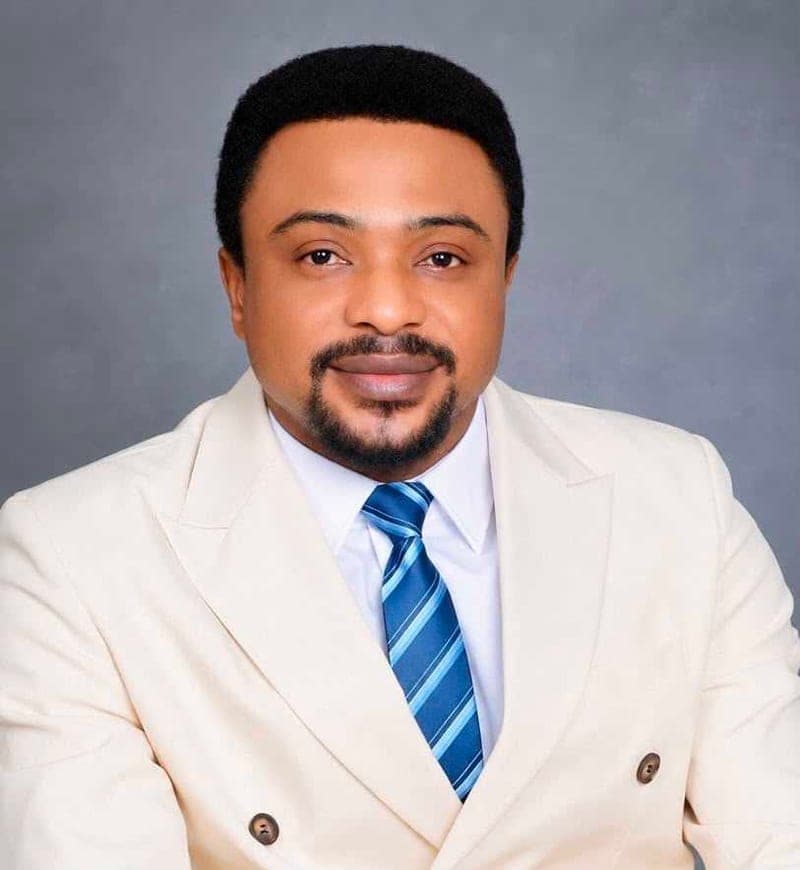 Pastor Ajonye Daniel Ajonye in a cream double-breasted suit and striped tie, against a grey studio backdrop